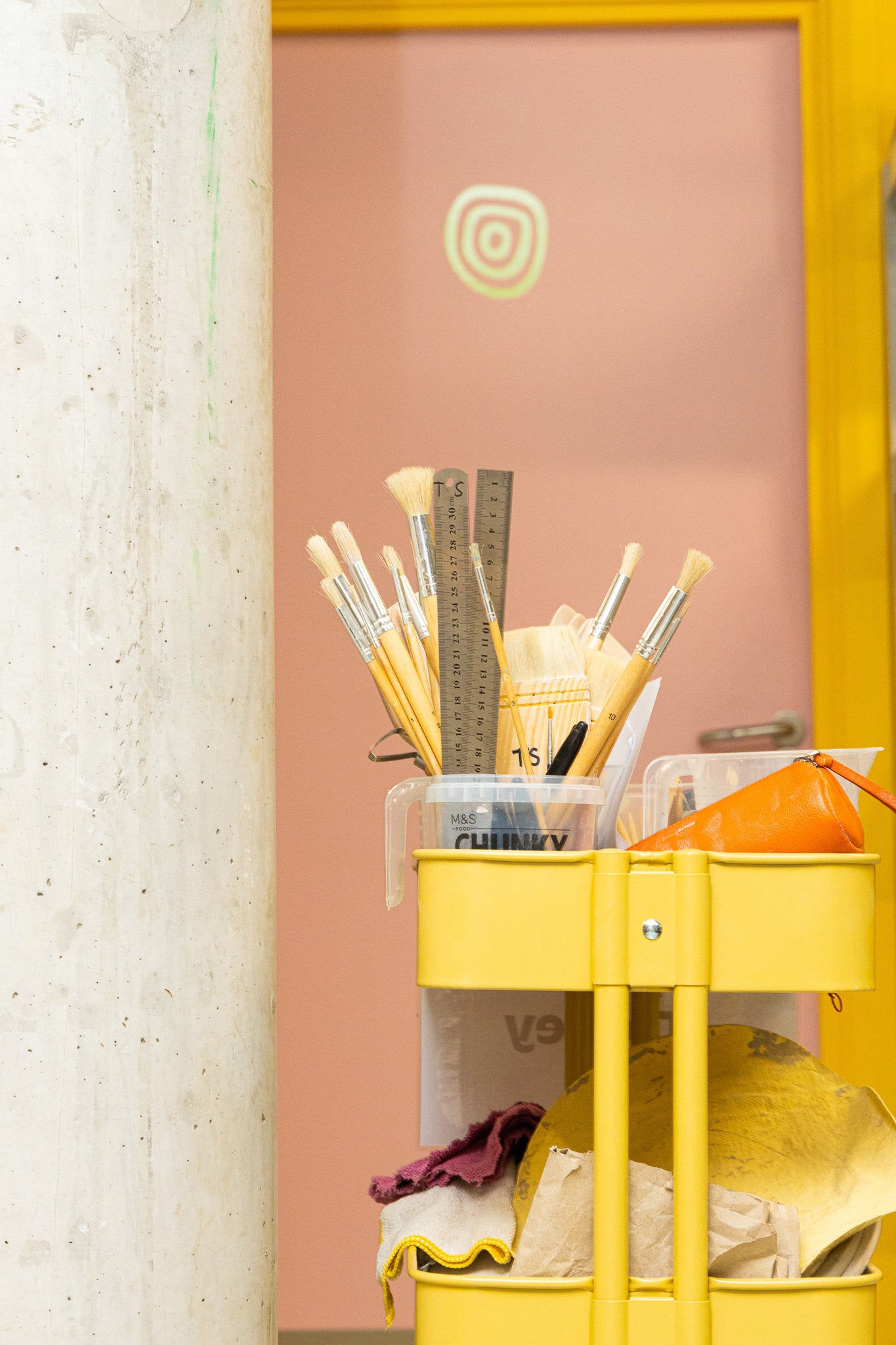 Paintbrushes and other hand-building tools are stacked on a bright yellow trolley in front of a salmon pink door. 