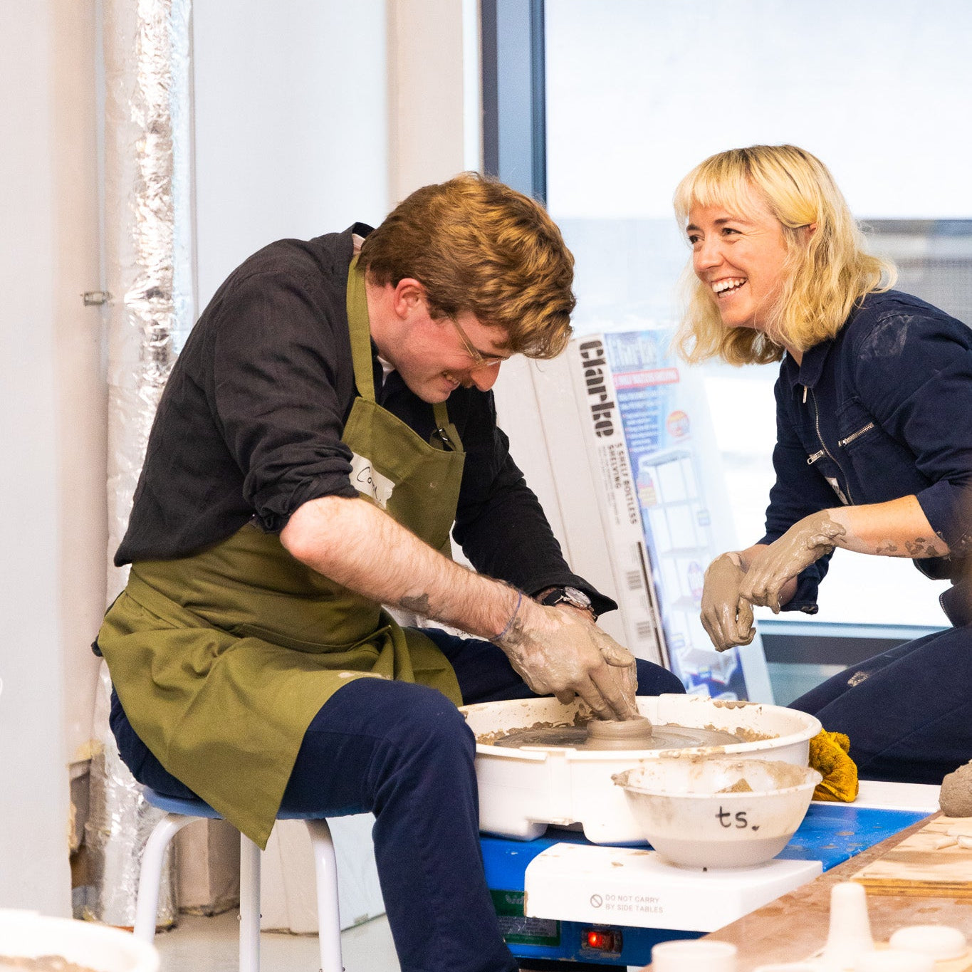 Throwing Shapes founder Siofra smiles whilst sitting beside a student at the potter's wheel whilst they practice.