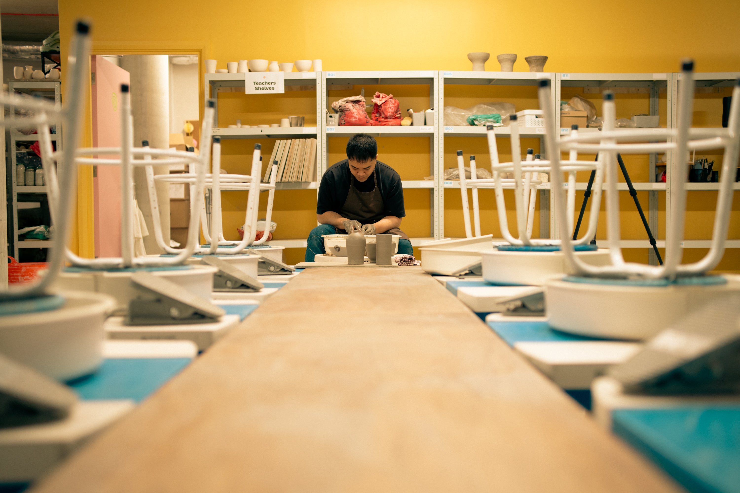 Throwing Shapes teacher Vinh sits at the potter's wheel deep in concentration. He sits at the end of a long wooden bench either side of which has a number of pottery wheels and stools.