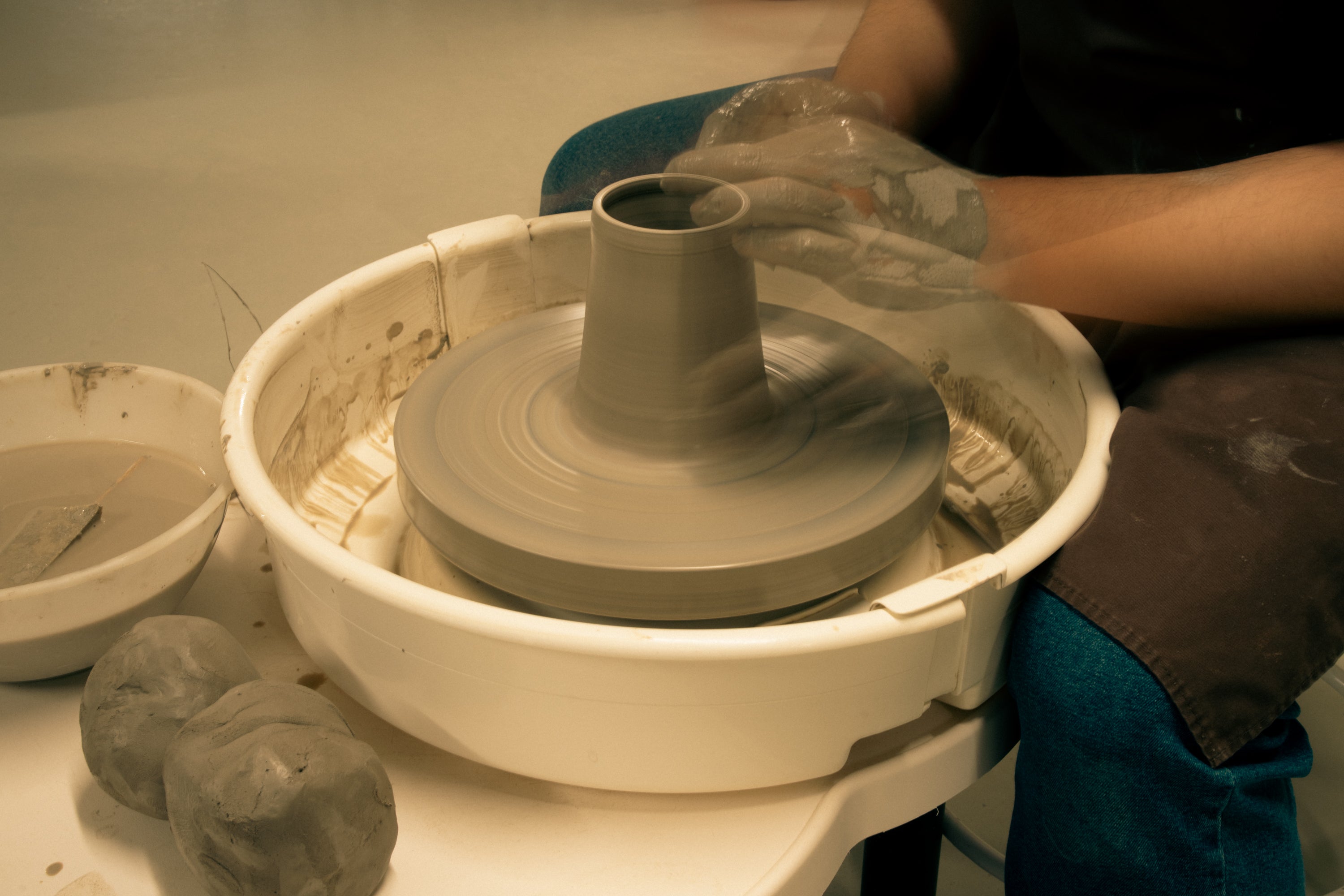 Throwing Shapes teacher Vinh sits at the potters wheel to throw a clay vase. The speed of the wheel is apparent from the photograph as motion blur can be seen. 