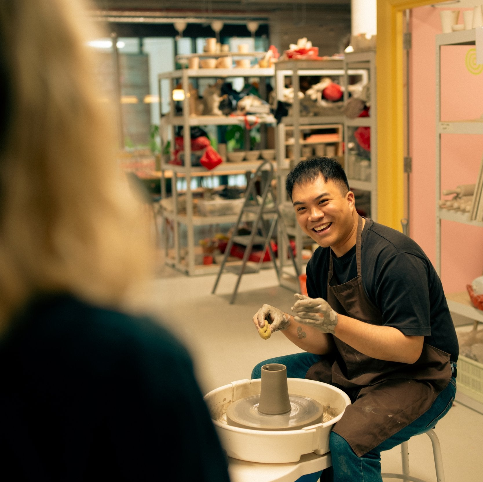 Throwing Shapes teacher Vinh smiles at studio founder Siofra whist sitting at the potter's wheel. A freshly made clay vase rests on the wheel in front of him.