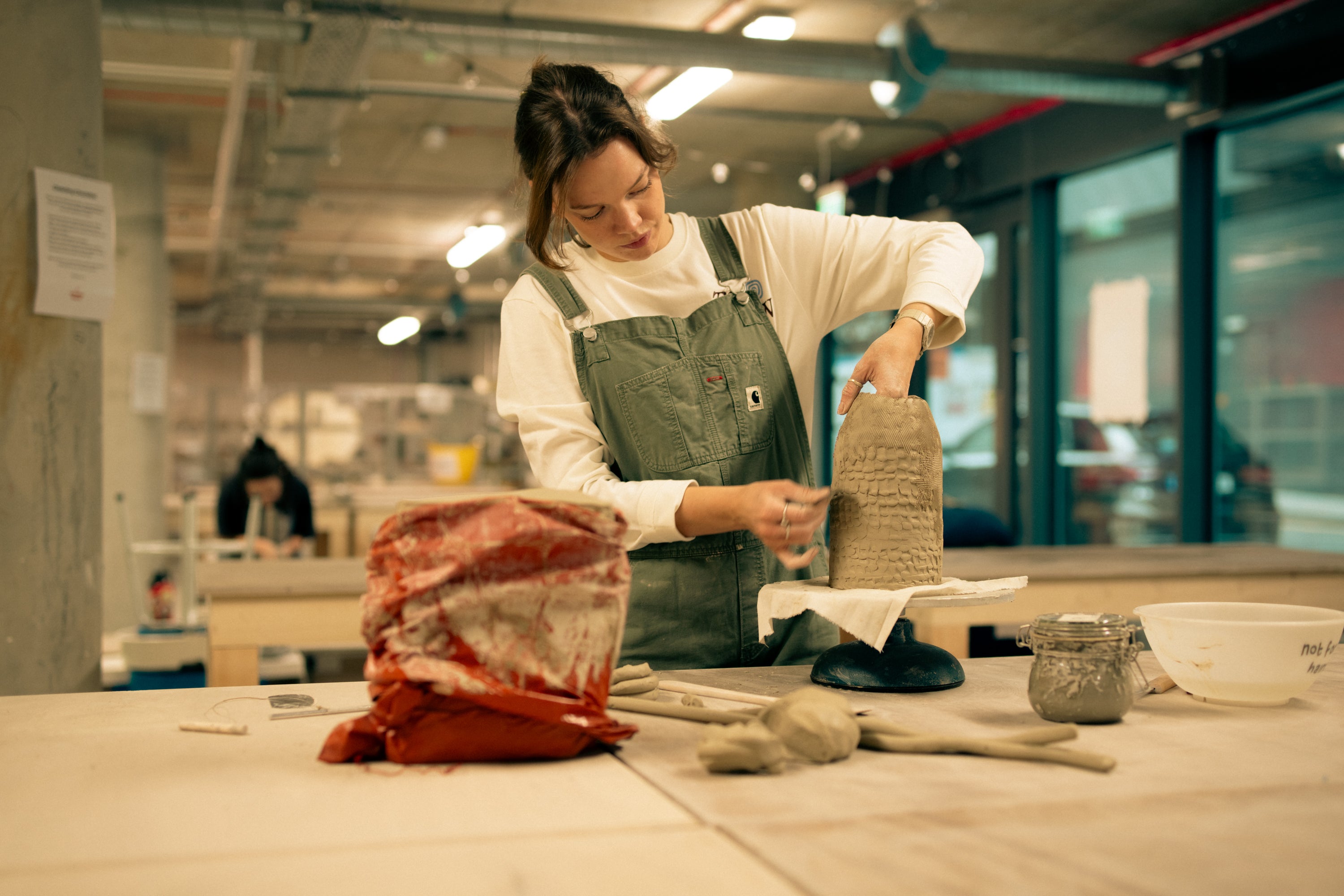 Throwing Shapes teacher Aine stands at a wooden bench whilst hand-building with clay. Tools and clay wrapped in plastic are shown at the workbench.