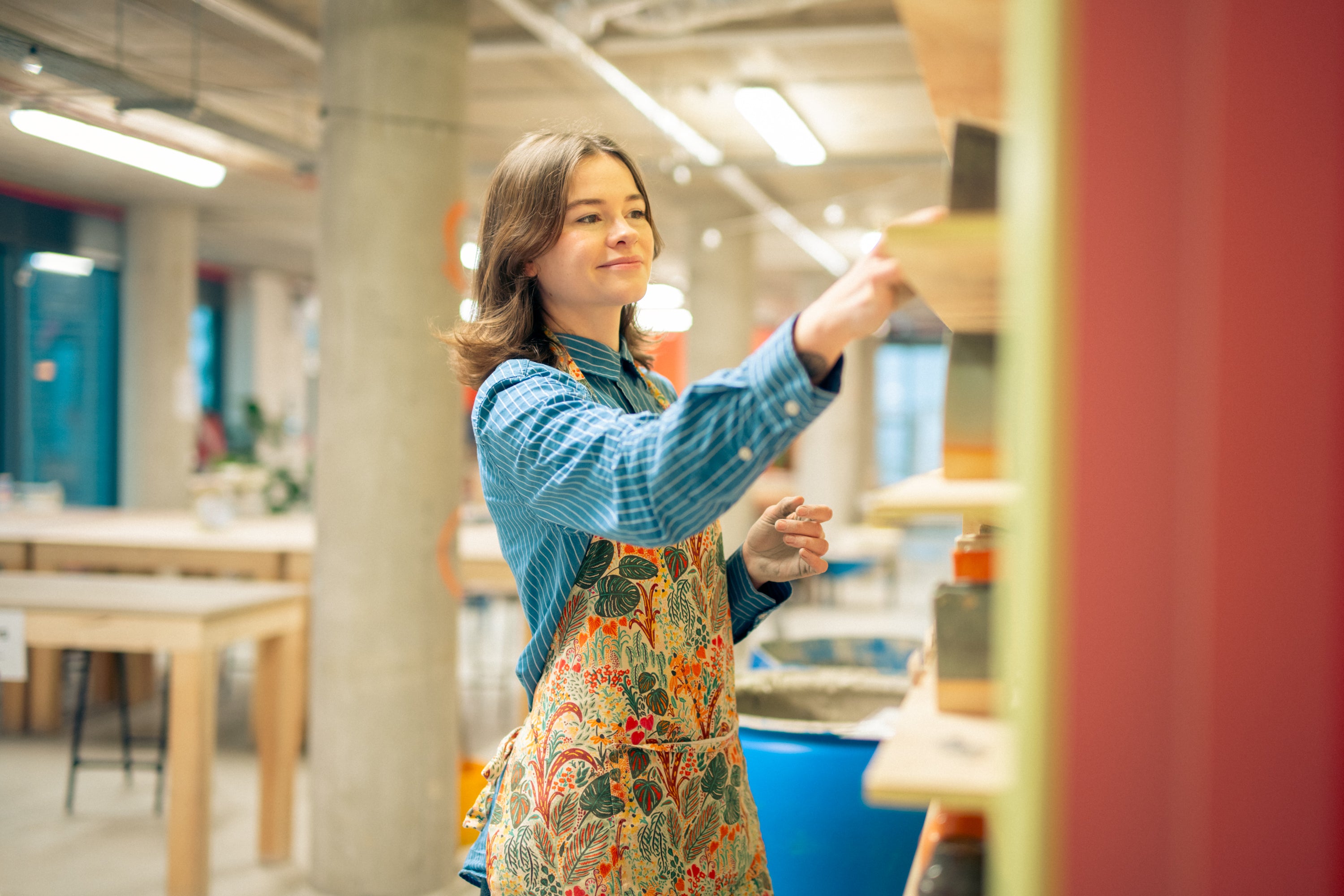 Throwing Shapes teacher Lily reaches out to the wall of glazes. Wooden benches can be seen behind her.