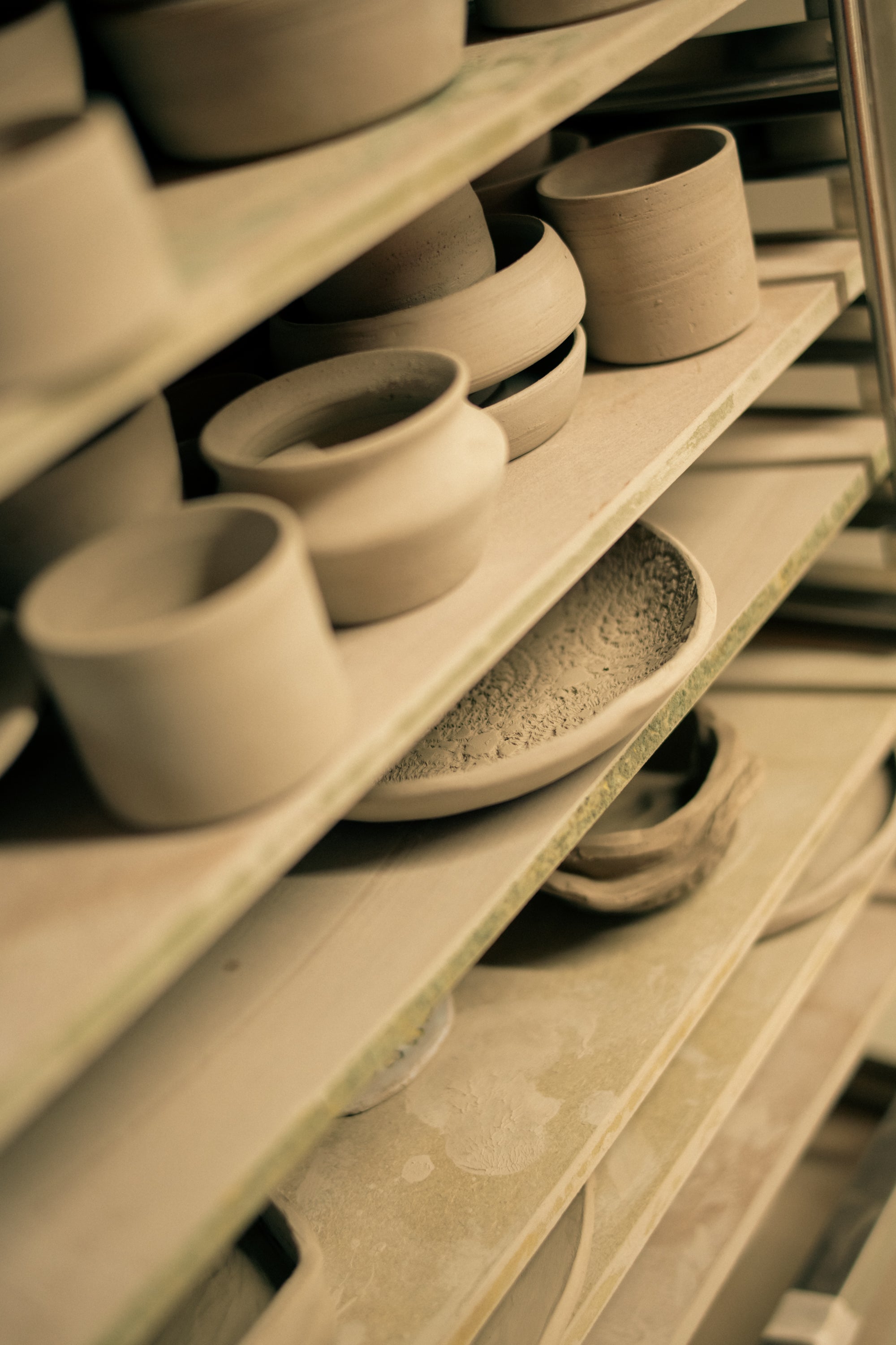 Multiple unglazed ceramic pieces rest on wooden shelves in preparation for glazing