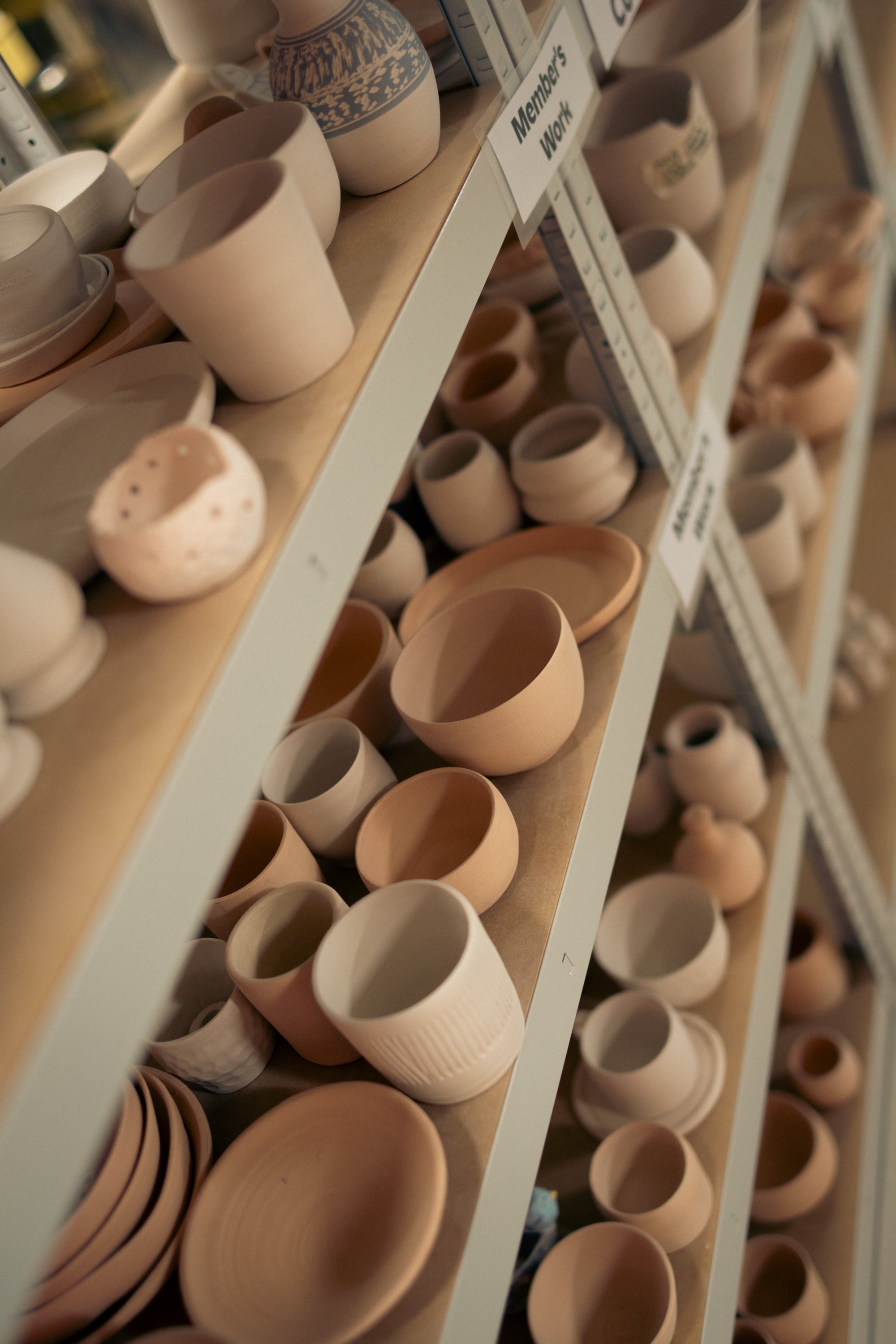 Multiple unglazed ceramic pieces rest on wooden shelves in preparation for glazing