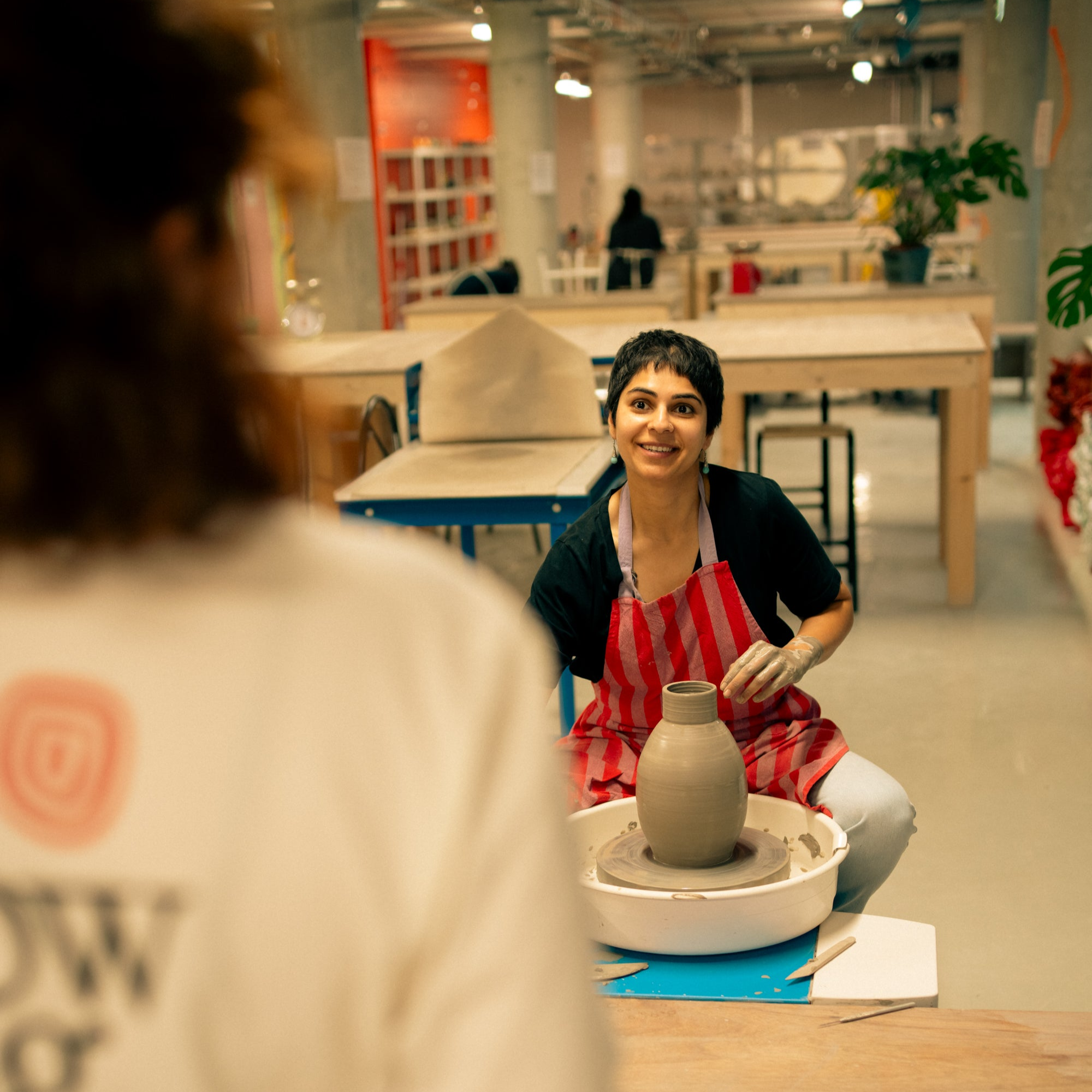 Throwing Shapes teacher Mahima smiles at studio manager Emma as she sits at the potter's wheel. A freshly made clay vase sits on the wheel in front of her.