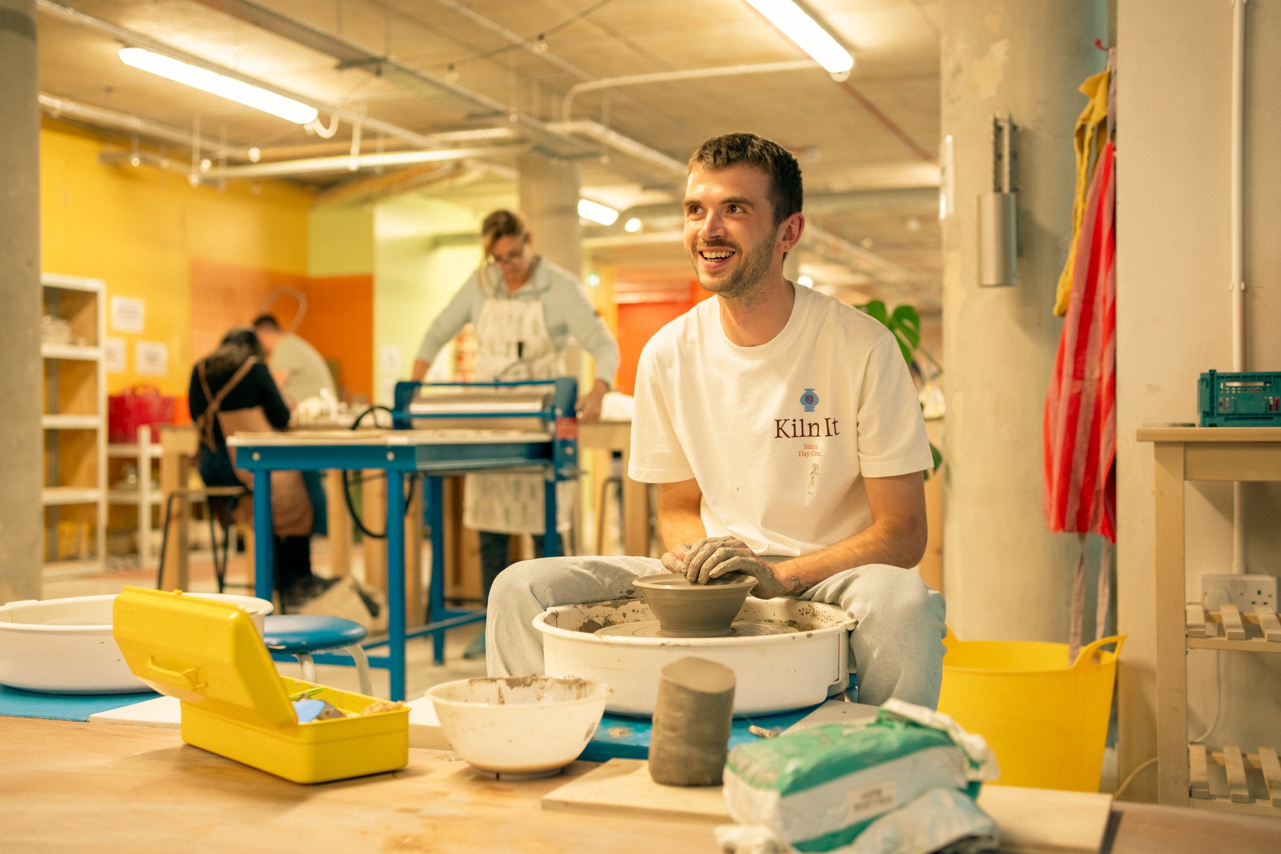 Throwing Shapes teacher Simon smiles as he sits at the potter's wheel throwing a small bowl. Tools and clay are to the forefront of the image. To the back studio three members are practising ceramics also. 