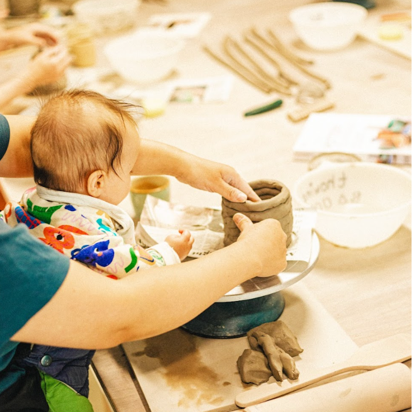 A baby in a sling reaches out towards a clay point their parent is building with coils. 