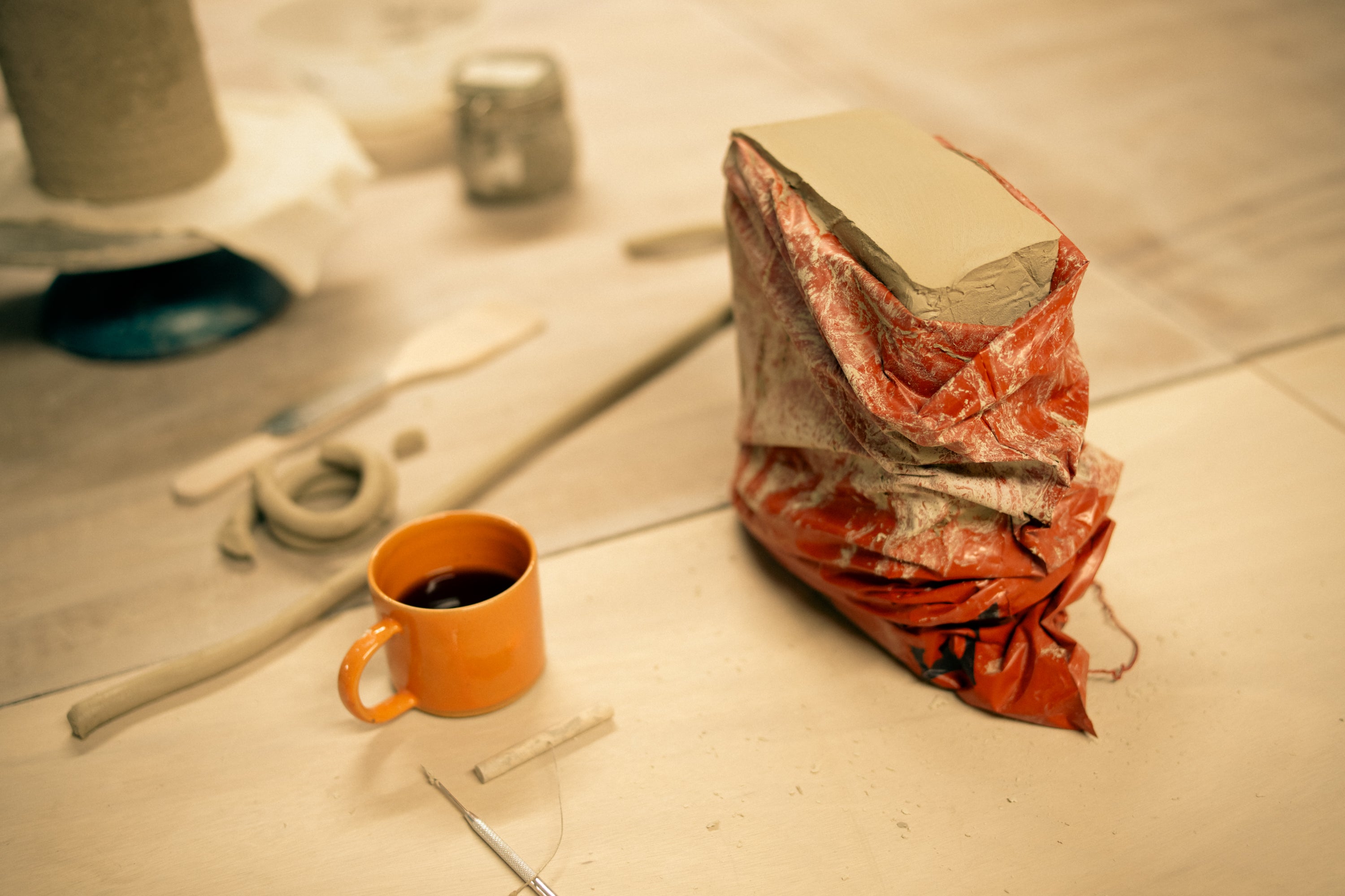 Clay wrapped in plastic sits on a wooden bench alongside a cup of black coffee and hand-building tools. 